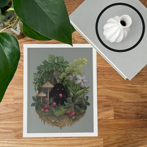 May include: A rectangular art print featuring a central illustration of a small black creature with glowing red eyes holding a red berry. The creature is surrounded by green foliage, mushrooms, and purple flowers on a gray background.