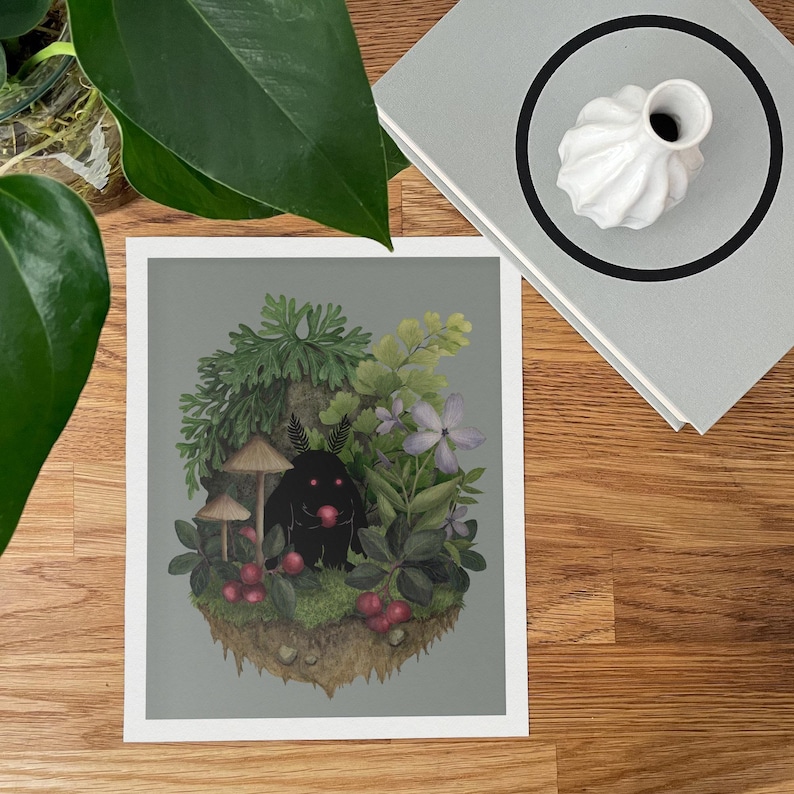 May include: A rectangular art print featuring a central illustration of a small black creature with glowing red eyes holding a red berry. The creature is surrounded by green foliage, mushrooms, and purple flowers on a gray background.