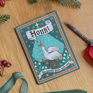 May include: A greeting card with a green background and a white goose illustration. The card reads "Honk!" in a banner and "The herald goose both sing" on a ribbon. A decorative pen rests beside the card.