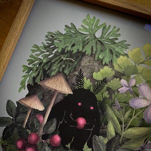 May include: A framed art print featuring a central black creature with glowing pink eyes holding a red berry. The creature is surrounded by two light brown mushrooms, dark green foliage, red berries, and light purple flowers on a gray background.
