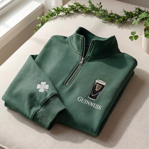 Op de afbeelding: Een donkergroene trui met een kwartrits, een wit geborduurd Guinness-logo en een klavertjevier op de mouw. De trui is opgevouwen op een neutrale ondergrond.