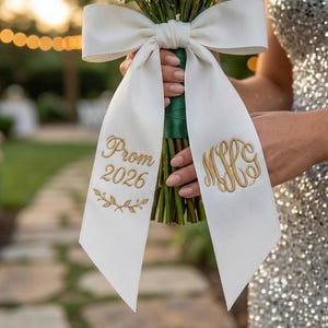 May include: A bouquet of yellow roses and baby's breath tied with a white ribbon. The ribbon is embroidered with "Prom 2026" and a monogram in gold thread. The bouquet is held by a person wearing a silver sequined dress.
