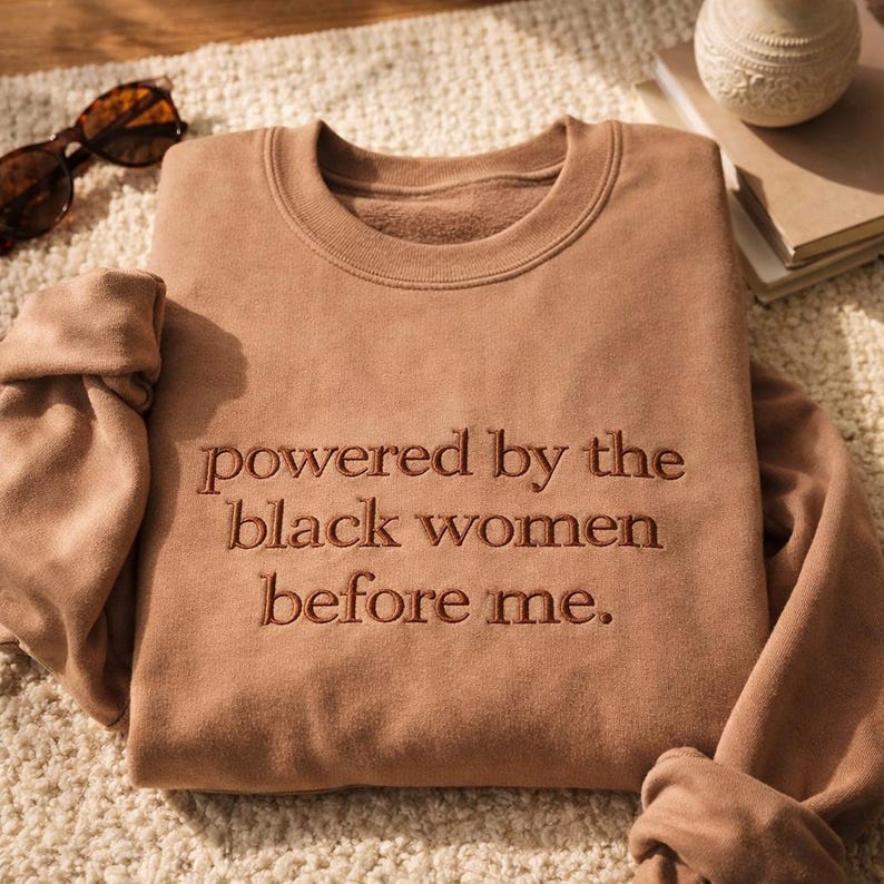 May include: A tan sweatshirt with the embroidered text "powered by the black women before me." The crew neck sweatshirt is folded on a textured surface, with sunglasses and a decorative vase in the background.