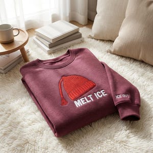 Puede incluir: Una sudadera granate con un diseño de gorro de punto rojo y las palabras "MELT ICE" y "ICE OUT" en blanco. La sudadera está sobre una alfombra blanca y esponjosa.