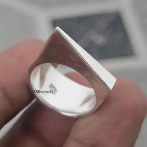 May include: A silver ring with a geometric design. The ring features a flat, rectangular top that slopes down to the band. The ring is highly polished and reflects light. The text "KSliverarts" is visible on the ring.