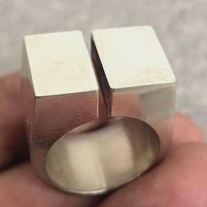 Puede incluir: Un anillo de plata con un diseño único. El anillo presenta dos bloques rectangulares unidos por una banda. El anillo tiene un estilo moderno y geométrico.