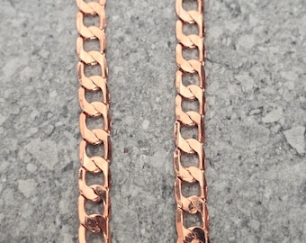 May include: A close-up shot of a copper-colored curb chain necklace. The chain links are thick and rectangular, with a polished finish. The necklace is displayed against a gray, textured background.