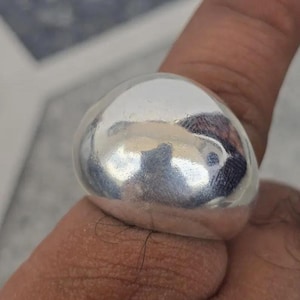 以下が含まれることがあります： 滑らかで丸みを帯びたドーム型の、大きく磨かれたシルバーリング。 リングは光を反射し、表面にハイライトと影を作り出しています。 リングは指に装着されています。