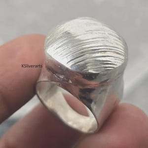 May include: A silver ring with a textured, domed top. The ring has a wide band and a smooth, polished interior. The surface of the ring is marked with horizontal lines. The text "KSliverarts" is visible.