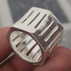 Könnte beinhalten: Ein silberfarbener Ring mit geometrischem Design. Der Ring hat eine rechteckige Form mit vertikalen Aussparungen um das Band. Der Ring wird in einer Nahaufnahme gezeigt.