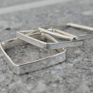 May include: Three silver square rings are stacked on a grey surface. The rings have a simple, minimalist design with a slightly hammered texture. The rings are made of a shiny, reflective metal.
