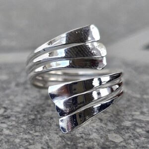 May include: A silver ring with a distinctive, layered design. The ring has multiple curved, overlapping bands, creating a flowing, sculptural look. The polished metal reflects light, highlighting the ring's intricate form.