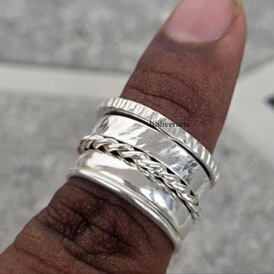 Puede incluir: Un anillo giratorio de plata con una banda texturizada y un diseño de cuerda trenzada. El anillo se lleva en un dedo. El anillo tiene un acabado brillante y pulido. Es una joya.