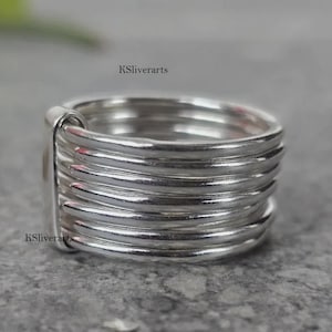 Puede incluir: Un anillo de plata con un diseño multicapa. El anillo presenta múltiples bandas de plata, creando un aspecto texturizado y dimensional. El anillo se muestra sobre una superficie gris. El texto "KSliverarts" es visible.