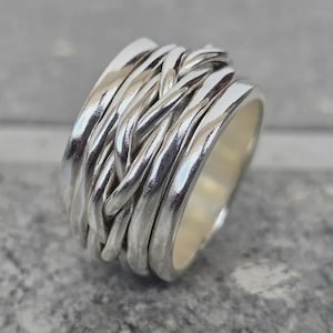 Puede incluir: Un anillo de plata con un diseño tejido. El anillo presenta múltiples bandas de metal pulido, creando una apariencia texturizada e intrincada. El diseño del anillo es una mezcla de elementos lisos y retorcidos.