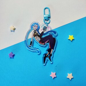 Può includere: Un ciondolo in acrilico trasparente con un personaggio con i capelli blu, che indossa un top nero e pantaloni viola. Il ciondolo ha un fermaglio blu e un'illustrazione a cartoni animati. Lo sfondo è blu con decorazioni a forma di stella.