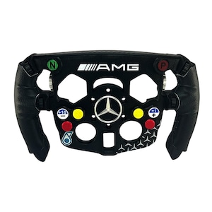 Puede incluir: Un volante de carreras negro con el logotipo AMG, un emblema central de estrella Mercedes-Benz y varios botones de colores etiquetados con N, P, +10, - y +1. Las empuñaduras son negras texturizadas y la sección inferior incluye un logo Petronas.
