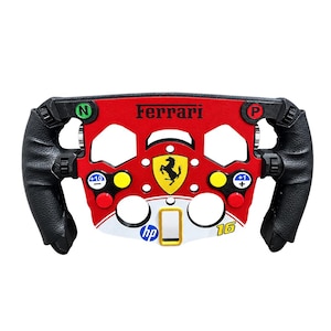 Könnte beinhalten: Ein Lenkrad im Formel-1-Stil mit einer rot-weißen Front, dem Ferrari-Logo, dem Schriftzug Ferrari und der Nummer 16. Es verfügt über schwarze, strukturierte Griffe, verschiedene farbige Tasten mit den Beschriftungen N, P, +10 und +1 sowie ein hp-Logo.