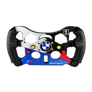 Pode incluir: Um volante de corrida BMW M4 Motorsport com estrutura preta e prateada, pegas em couro preto texturizado e um logótipo central da BMW. O volante inclui botões vermelhos e amarelos, um ícone quadrado com o número 1 e o texto M4 Motorsport.