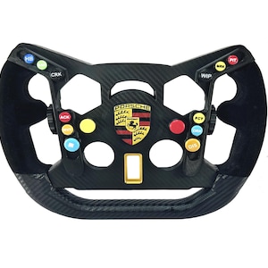 Può includere: Un volante da corsa nero e in fibra di carbonio con più pulsanti colorati e il logo Porsche. Il volante ha una forma rettangolare con un mozzo centrale e un'area di presa rivestita in pelle scamosciata nera.