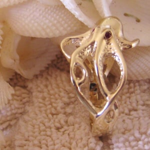 May include: A gold octopus ring with a red gemstone in the center. The ring is detailed with a textured surface and is set against a background of white seashells.