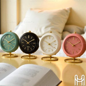 Puede incluir: Cuatro pequeños relojes de cuarzo redondos en colores pastel: verde azulado, negro, blanco y rosa. Cada reloj tiene una base y manecillas doradas. Las esferas muestran la palabra "QUARTZ".