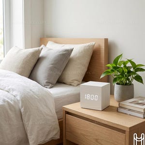May include: A wooden cube digital clock displaying 18:00, resting on a wooden nightstand. The bedroom setting includes a bed with pillows and a small potted plant. The clock is white with a digital display.