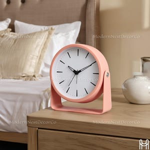 May include: A round, peach-colored analog clock with a white face and black hands and markers. The clock has a stand and is sitting on a wooden nightstand. The clock is a decorative item.