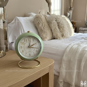 May include: A mint green analog alarm clock with a gold base and accents. The clock face is white with black numerals and gold hands. The clock is sitting on a light wood nightstand. The background includes a bed with white linens and fluffy pillows.