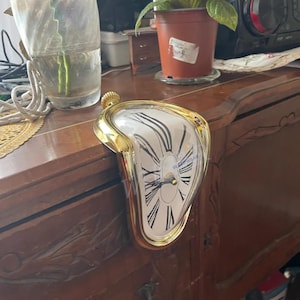 Może przedstawiać: Złoty, topiący się zegar z białą tarczą i czarnymi rzymskimi cyframi. Zegar ma unikalny, surrealistyczny design, z tarczą, która wydaje się kapać. Zegar jest przedmiotem dekoracyjnym.