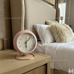 Puede incluir: Un reloj despertador analógico rosa con una base dorada está sobre una mesita de noche de madera. La esfera del reloj tiene números negros y manecillas doradas. El fondo incluye una cama con ropa de cama blanca y un cabecero beige. El reloj tiene unos 10 cm de diámetro.
