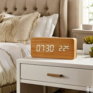 May include: A rectangular wooden digital alarm clock displaying the time as 07:30 and the temperature as 22 degrees Celsius. The clock has a light wood grain finish and is placed on a white nightstand.