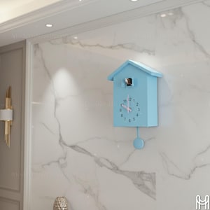Op de afbeelding: Een lichtblauwe koekoeksklok in de vorm van een huis, aan een muur met marmerpatroon. De klok heeft een ronde slinger en een kleine vogel zichtbaar in het raam. De wijzers zijn roze.
