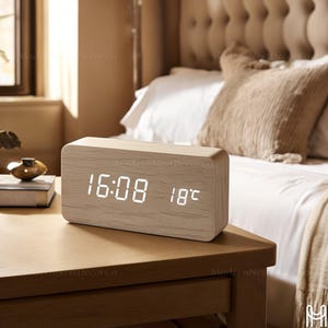 Può includere: Un orologio digitale rettangolare in legno che mostra le 16:08 e 18°C. L'orologio, con finitura a venatura di legno chiaro, è posto su un comodino in legno. Lo sfondo include un letto con lenzuola bianche e una coperta beige.