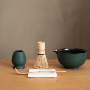 May include: A matcha tea set featuring a dark teal ceramic bowl, a bamboo whisk, a small teal ceramic cup, a bamboo scoop, and a white cloth, all arranged on a wooden surface. The bowl has a black interior and a spout.