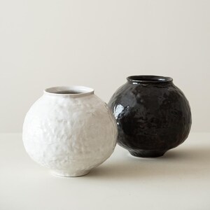 May include: Two ceramic vases. One is white with a textured surface, the other is black and glossy. Both have a round body and a narrow opening at the top. The vases are displayed against a neutral background.