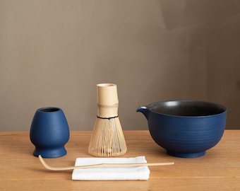 May include: A matcha tea set featuring a navy blue bowl, a whisk, a small blue cup, a bamboo scoop, and a white cloth. The bowl has a dark interior and a spout. The whisk is made of bamboo. The set is arranged on a wooden surface.
