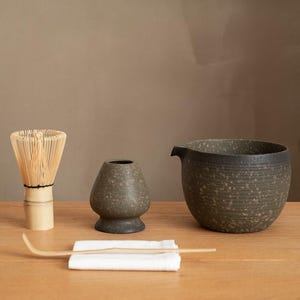 May include: A matcha tea set featuring a bamboo whisk, a small ceramic container, a tea bowl with a spout, a bamboo scoop, and a white cloth. The tea set is made of natural materials and has a minimalist aesthetic.