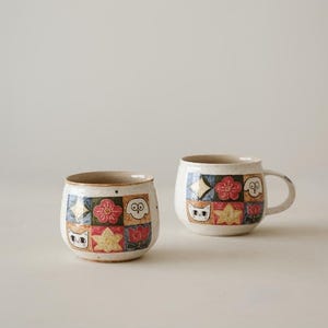 May include: Two ceramic mugs with a speckled off-white base, each featuring a grid pattern of colorful squares. The squares contain hand-painted illustrations of a cat face, a dog face, a red flower, a yellow star, and geometric shapes.