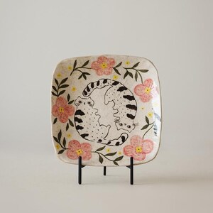 May include: A square ceramic plate with rounded corners, featuring a hand-painted design of two cats in a circle, surrounded by pink flowers and green leaves. The plate is displayed on a black metal stand, showcasing a whimsical, floral-themed design.