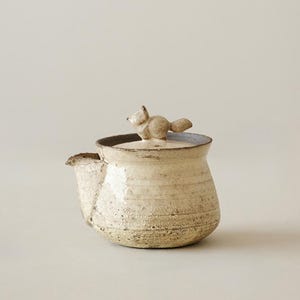 May include: A small, off-white ceramic teapot with a textured, speckled finish. The lid features a sculpted, three-dimensional fox figure acting as a handle. The teapot has a wide, rounded body and a short, integrated spout on the left side.