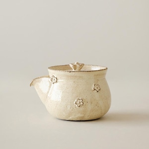 May include: A cream-colored ceramic teapot with a speckled finish and a rounded body. The teapot features a small spout, a matching lid with a flower-shaped knob, and several small, embossed flower motifs scattered across the exterior surface.