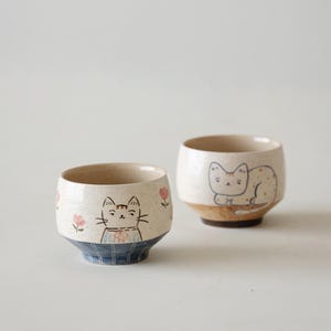 May include: Two ceramic cups with a speckled cream finish. The front cup features a painted cat face with pink flowers. The back cup displays a cat resting on a brown base. Both cups have a rounded shape and a distinct bottom section with blue patterns.