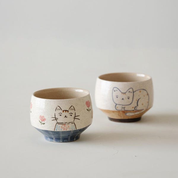 May include: Two ceramic cups with a speckled cream finish. The front cup features a painted cat face with pink flowers. The back cup displays a cat resting on a brown base. Both cups have a rounded shape and a distinct bottom section with blue patterns.