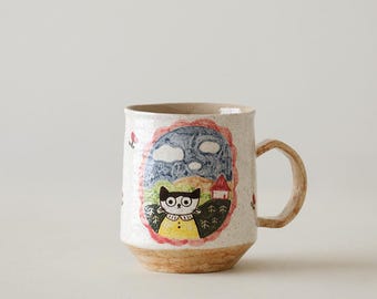 May include: A ceramic mug with a whimsical illustration of a cat-like figure in a landscape. The mug features a beige base and handle, with a colorful design of a cat, house, and trees. The mug is decorated with small red flowers.