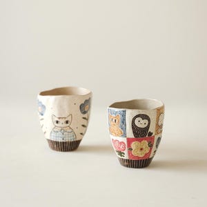 May include: Two ceramic cups with unique designs. One cup features a cat illustration with blue and white accents. The other cup showcases a patchwork design with various animal and floral motifs in multiple colors. Both cups have a textured, off-white base.