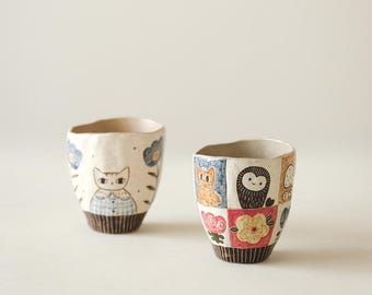 May include: Two ceramic cups with unique designs. One cup features a cat illustration with blue and white accents. The other cup showcases a patchwork design with various animal and floral motifs in multiple colors. Both cups have a textured, off-white base.