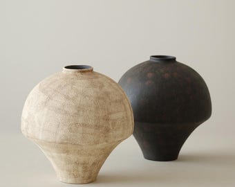 May include: Two ceramic vases. One is a light beige color with a textured surface, and the other is dark brown with a speckled pattern. Both have a rounded body and a narrow neck, with a flared base. The vases are displayed on a neutral background.