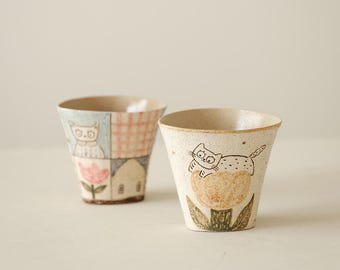 May include: Two ceramic cups with hand-drawn illustrations. One cup features a cat on a flower, the other has a patchwork design with a cat, house, and flower. The cups have a beige base and are ideal for beverages.