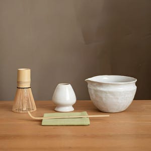 May include: A matcha tea set on a wooden surface. The set includes a bamboo whisk, a small white ceramic vase, a white ceramic bowl, a green cloth, and a bamboo scoop. The background is a solid brown color.